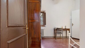 Un pasillo con puerta de madera, un radiador, una silla, una mesita y una cama con estructura metálica blanca.