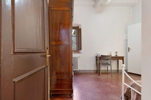 Un pasillo con puerta de madera, un radiador, una silla, una mesita y una cama con estructura metálica blanca.