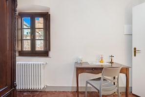 Une pièce comprenant un bureau en bois, une chaise, un radiateur, une fenêtre donnant sur des immeubles et une porte.