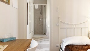 Habitación de hotel con una mesa de madera, una silla blanca, una cama con cabecera marrón y zona de ducha con puerta de vidrio.