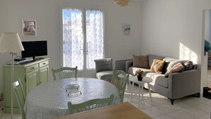 A living room with a dining table, a sofa, and a TV. There are two windows with curtains, a lamp, and a ceiling light.
