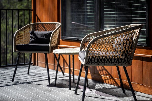 Two wicker chairs with cushions on a wooden deck, a small round table, and a window with blinds.