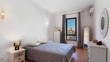 A bedroom with a bed, bedside table, lamp, dresser, mirror, and a window with a view.