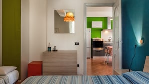 Une chambre à coucher comprenant un lit, une commode, un miroir et donnant sur un coin cuisine.