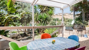 Une terrasse extérieure couverte, équipée d’une table, de chaises et offrant une vue sur la verdure et les bâtiments environnants.