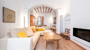 Una sala de estar con sofá, mesa de centro de madera y sillas. Se ve una chimenea, una mesa de comedor y una zona de cocina a través de un arco.