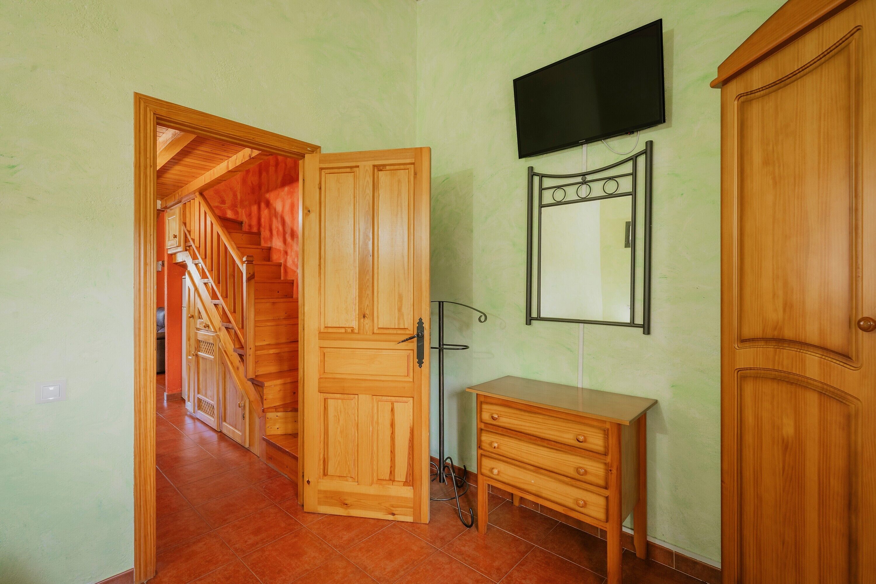 Una habitación con un armario de madera, un pequeño tocador de madera, un espejo y un televisor en la pared.