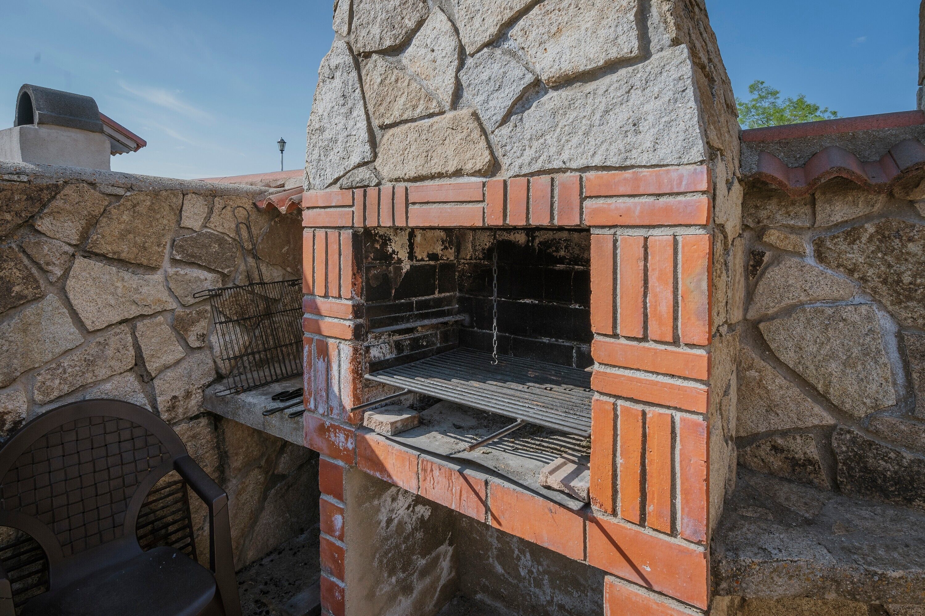 Un barbecue extérieur en pierre et en briques, doté d’une cheminée, d’une grille en métal et d’une chaise en métal.