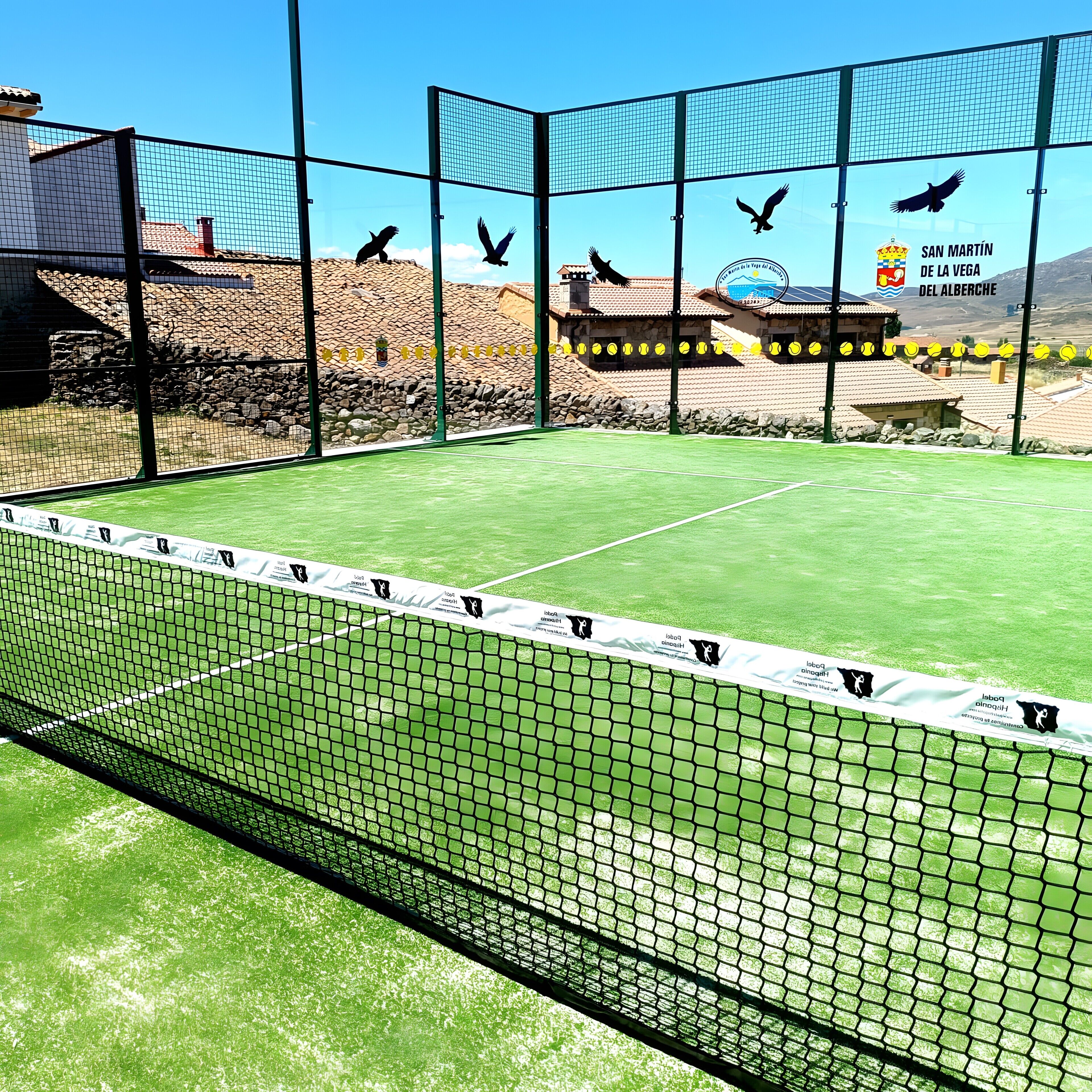 Una cancha de tenis con una red, rodeada por una cerca, y un edificio con un cartel que dice 'SAN MARTÍN DE LA VEGA DEL ALBERCHE' al fondo.