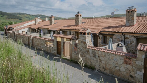 A row of stone houses with tiled roofs, a gated entrance, and a view of green hills in the background.