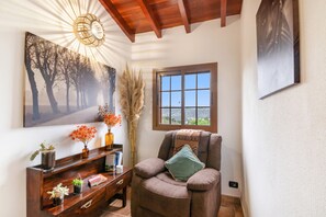 Ein Zimmer mit Holzboden, einem Fenster mit Blick, einem Sessel mit Kissen, einem Regal mit Pflanzen und einer Wandlampe.