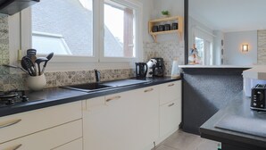 A modern kitchen with a black countertop, white cabinets, a window, and a shelf with kitchen items. - Holiday Home 'Maison Au Port De Lézardrieux' with Private Garden, Balcony and Wi-Fi (Lézardrieux)