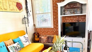 Una sala de estar con un sofá amarillo, una mesa redonda de centro con fruta y una caja de rosquillas, una guitarra y un televisor sobre una pared de ladrillo.