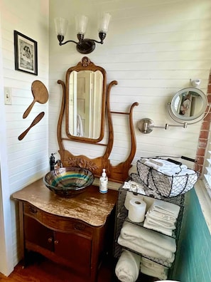Un baño con tocador de madera, un espejo grande, lavabo tipo cuenco, toallero y un estante pequeño con artículos de aseo.