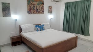 A bedroom with a wooden bed, white bedding, a nightstand, a wall-mounted air conditioner, and a painting above the bed. - beach resort casa (Sekondi-Takoradi)