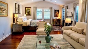 A well-lit room with a bed, a sofa, a chair, and a coffee table. There is a ceiling fan, a window with blinds, and a painting on the wall.