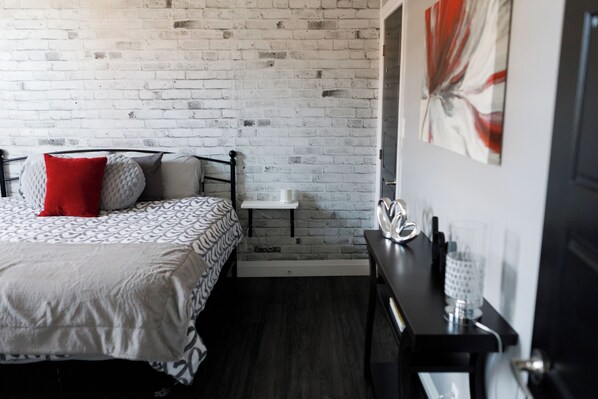 A bedroom with a brick wall, a bed with patterned bedding, a bedside table, and a wall-mounted artwork.