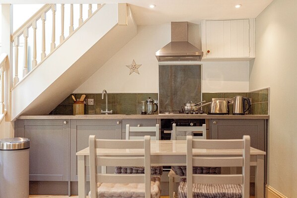 A compact kitchen with a built-in stove, a range hood, and a small dining area with a table and chairs. - Mill House & Cottage - on Cotswold Way (Stroud)