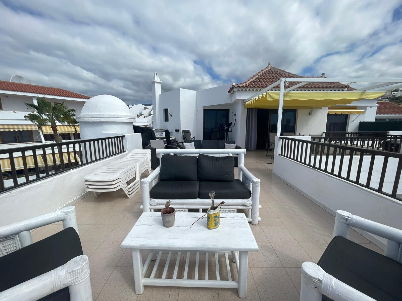 A rooftop terrace with white furniture, including a sofa and chairs, a small table with potted plants, and a yellow umbrella.