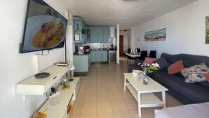 Una sala moderna con un sofá, una mesa de centro y un televisor. Se ve una cocina al fondo con armarios azul claro y un comedor.