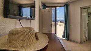 Una habitación con un televisor montado en la pared, un sombrero de paja sobre una mesa y una puerta que da a un balcón con vista al mar.