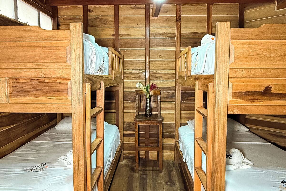 A wooden cabin interior with bunk beds, a small table with a vase, and a patterned rug.