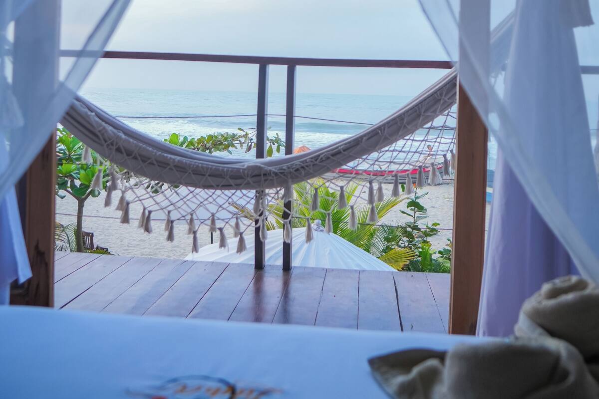 A hammock on a wooden deck overlooking a beach with a view of the ocean.