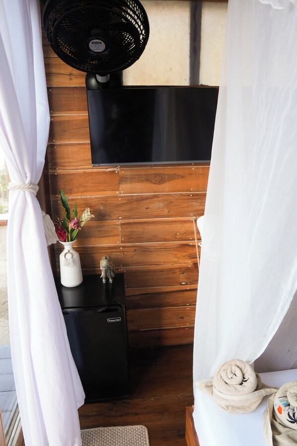 A room with a wooden wall, a black fan, a small white table with a vase of flowers, and a TV mounted above the table. - Luma at the beach (Santa Marta)