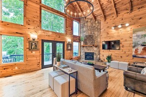 Una cabaña de madera espaciosa con una amplia sala de estar que incluye chimenea, asientos cómodos y ventanas grandes con vistas al bosque circundante.