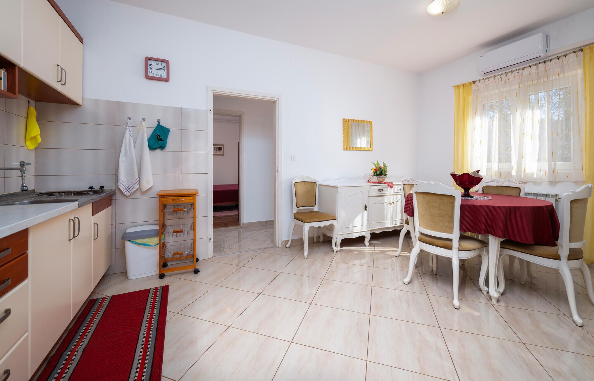 Una cucina compatta con lavandino, piano cottura e pensili, una zona pranzo con tavolo e sedie, e un piccolo corridoio che conduce a una camera da letto.