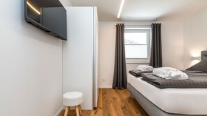 Ein modernes Schlafzimmer mit einem Bett, einem kleinen Hocker, einem an der Wand befestigten Fernseher und einem Fenster mit Vorhängen.