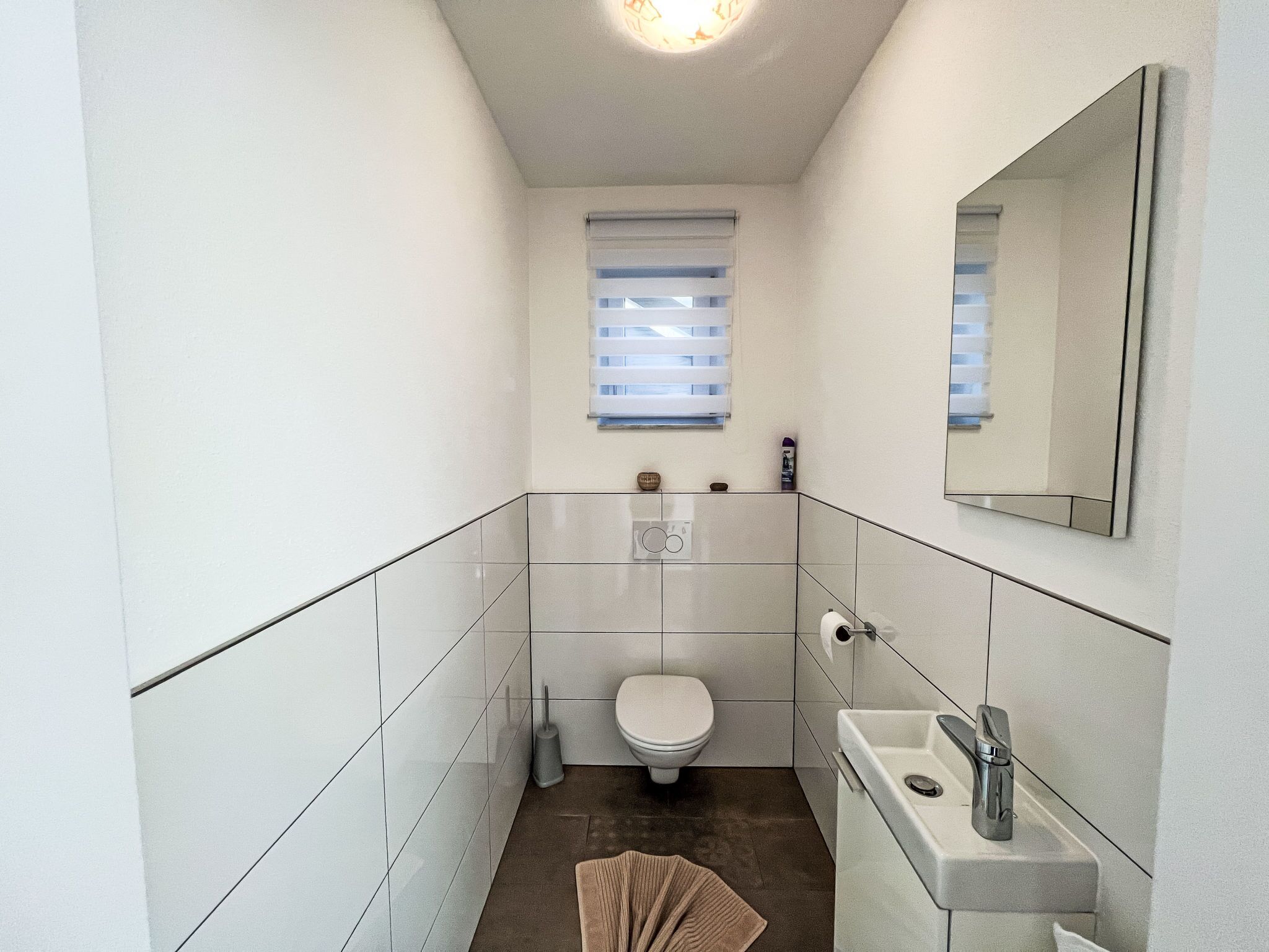 Ein kleines, modernes Badezimmer mit weißen Fliesen, einer Toilette, einem Waschbecken und einem Spiegel.