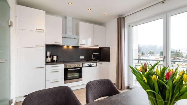 Une cuisine moderne avec des meubles blancs, un plan de travail noir et un four. On y trouve une table à manger avec deux chaises et un vase de tulipes.