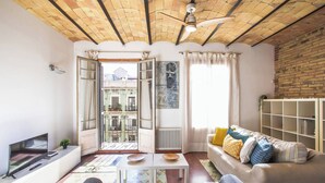 A living room with a sofa, a coffee table, a TV, and a bookshelf. There is a balcony with a view of buildings and a door leading outside.