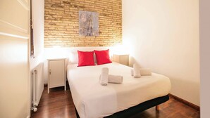 A bedroom with a brick wall, a bed with white bedding and red pillows, a nightstand, and a radiator.