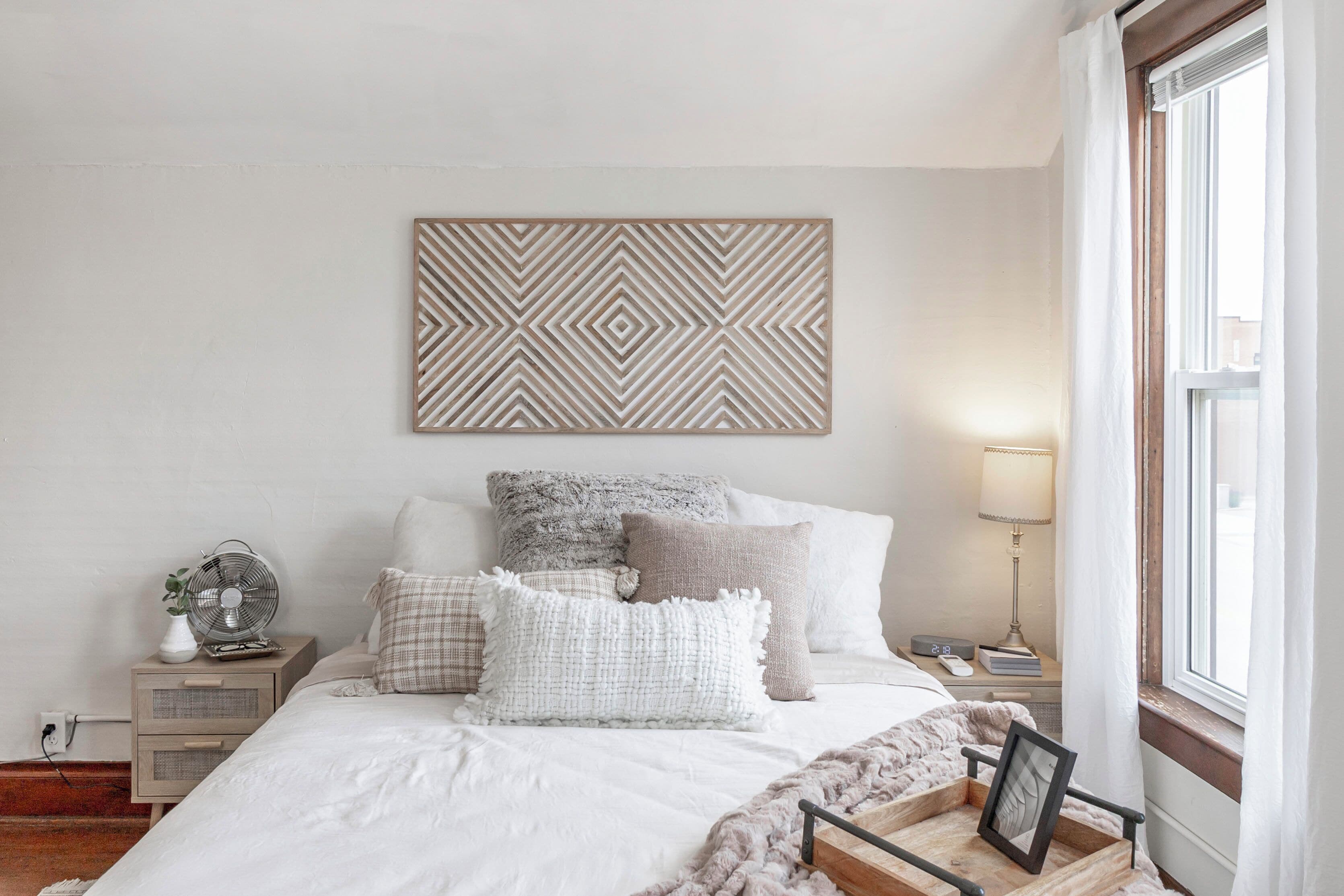 Un dormitorio ordenado con cama, almohadas, mesita de noche con lámpara y una obra de arte en la pared.