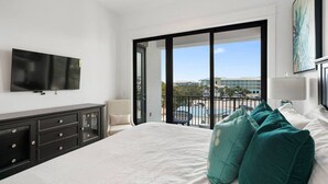 A hotel room with a large bed, a TV mounted on the wall, a balcony with a view of buildings, and a desk with drawers. - Get Busy Livin! on 30A (Santa Rosa Beach)
