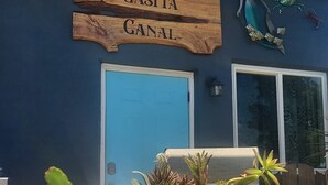 Un letrero de madera con la inscripción 'CASITA CANAL' sobre una puerta azul, con una silla y plantas en macetas al frente.