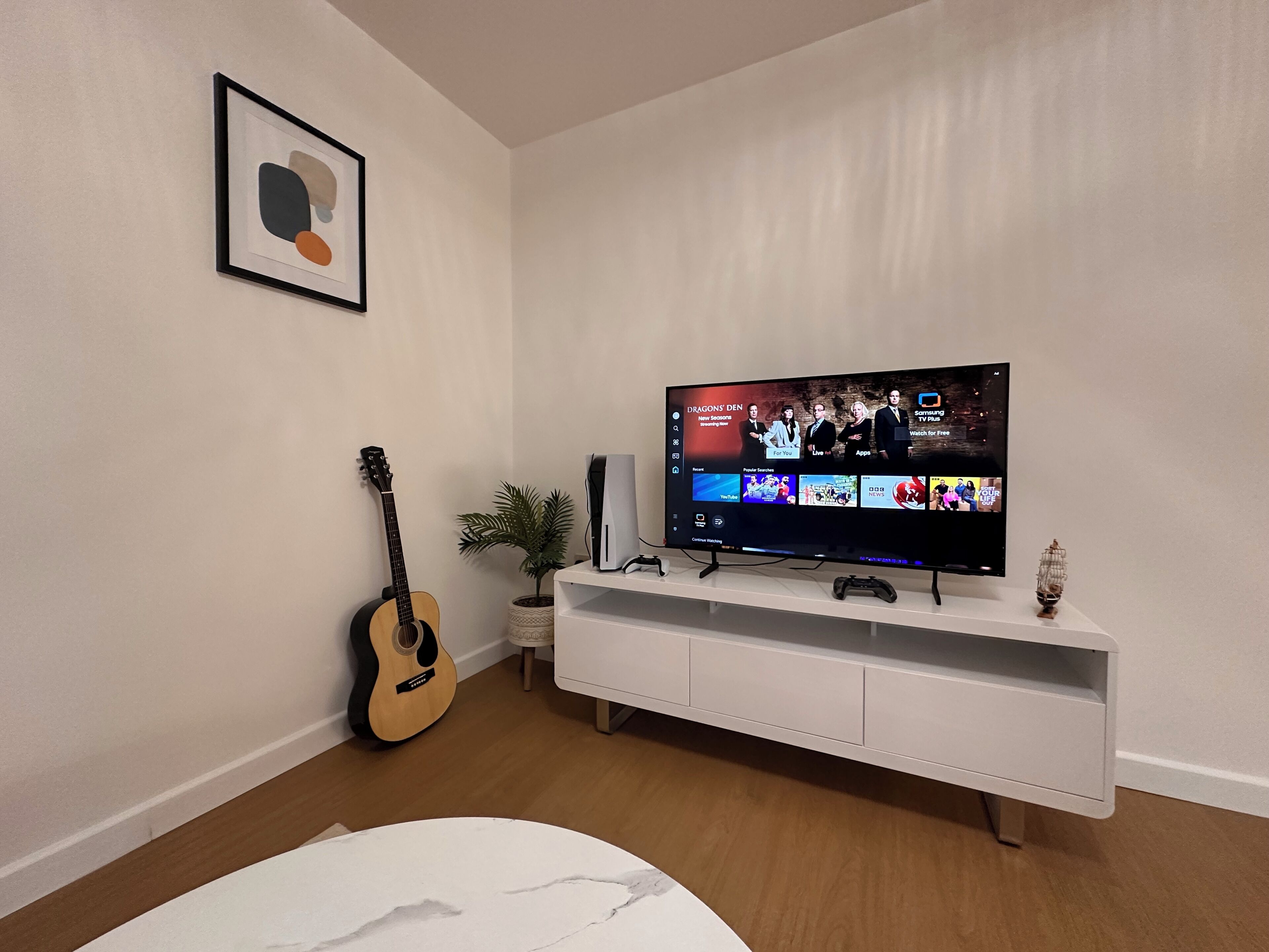 Un salón con televisor de pantalla plana sobre un mueble blanco, una guitarra, una planta en maceta y un cuadro en la pared.