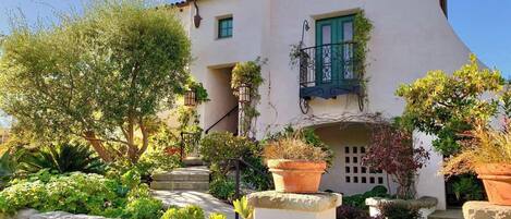 A white building with a red roof, a balcony with green railings, and a garden with various plants and a stone pathway.