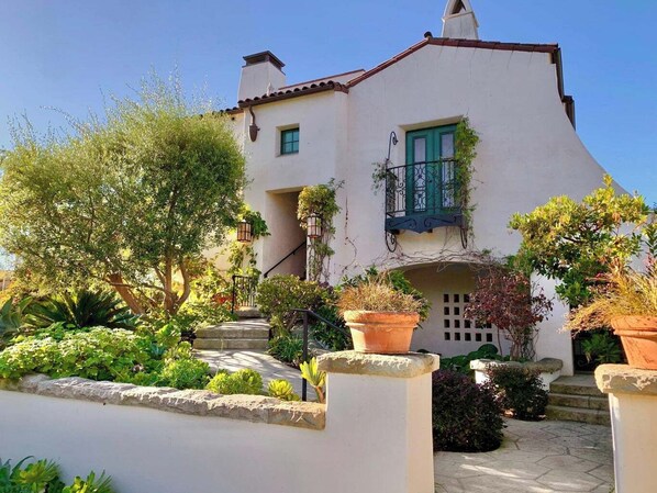 A white building with a red roof, a balcony with green railings, and a garden with various plants and a stone pathway.