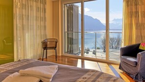 A bedroom with a large window, a bed, a chair, and a view of a lake and mountains.