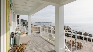 A covered outdoor area with a view of the beach and ocean. It includes a seating area with chairs and a table, a railing, and a staircase leading to an upper level. - Oceanfront Cottage with Private Beach Area! (Carolina Beach)