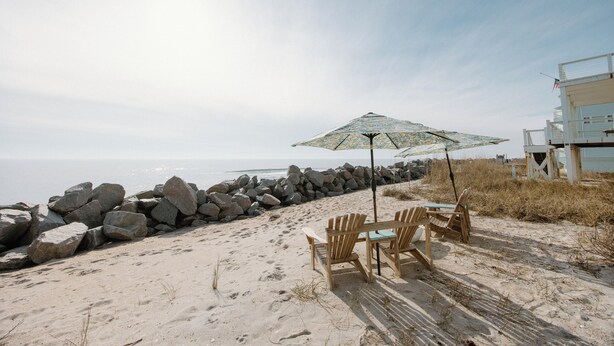 A beach with a wooden table and chairs under a beach umbrella, a rocky shoreline, and a lifeguard tower in the background. - Oceanfront Cottage with Private Beach Area! (Carolina Beach)