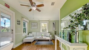 A living room with a white sofa, a coffee table, and a fireplace. There is a ceiling fan, a window, and a door with an exit sign. - Uptown Classic 1 | Effortless Exploration of STA (St. Augustine)