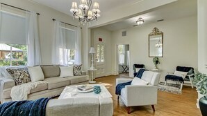 A spacious living room with a beige sofa, a white armchair, and a coffee table. There is a chandelier, a mirror, and a potted plant.