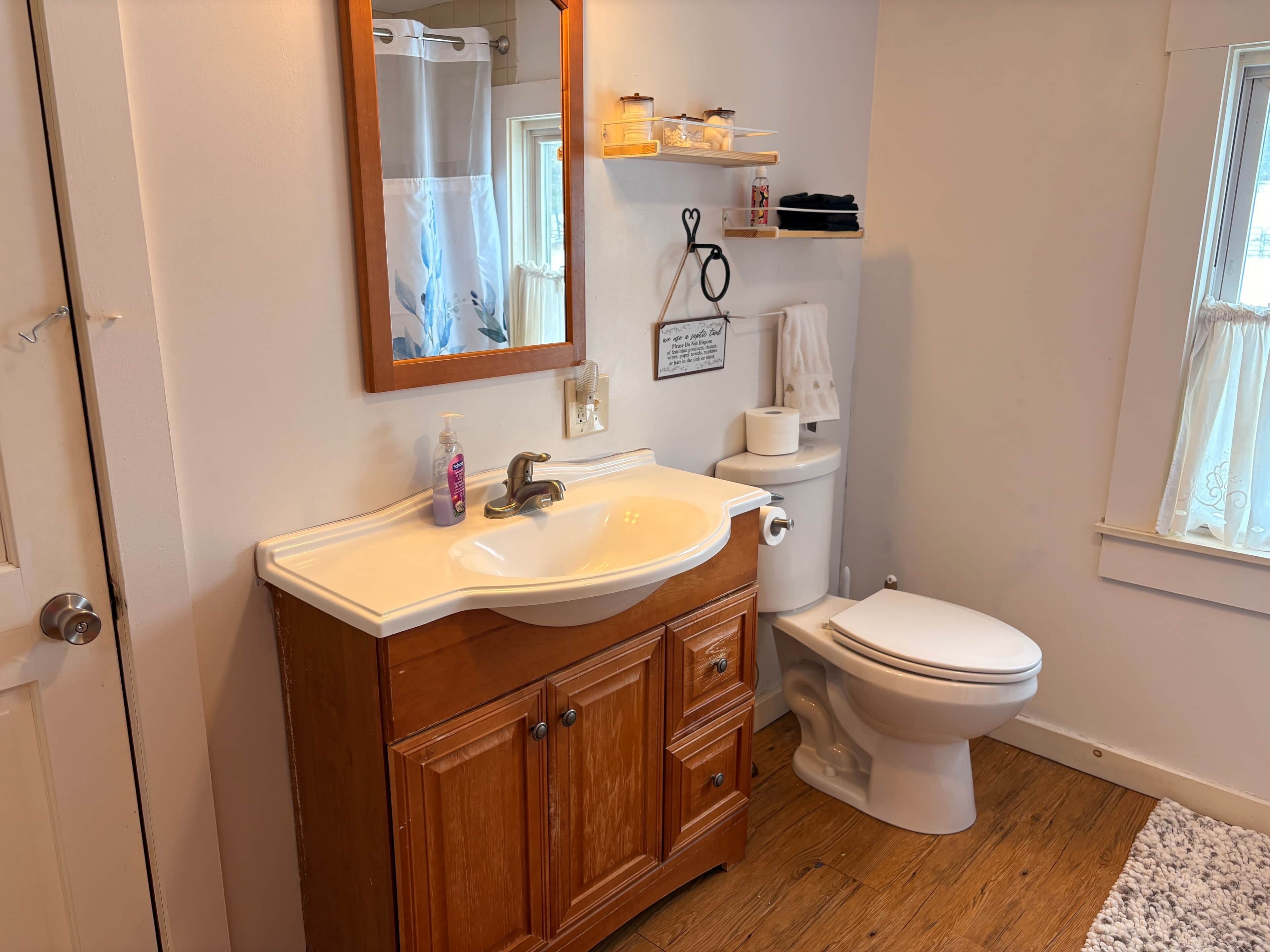 A bathroom with a wooden vanity, a white sink, a mirror, a toilet, and a window with a view.