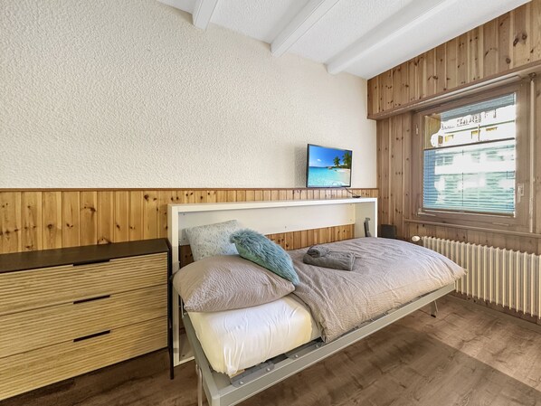 Quarto com cama, mesa de cabeceira, cabeceira de madeira, televisão na parede e janela com persianas.
