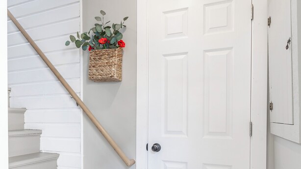 Un escalier aux marches blanches menant à une porte blanche, un petit tabouret et un panier rempli de fleurs rouges, le tout placé contre le mur.