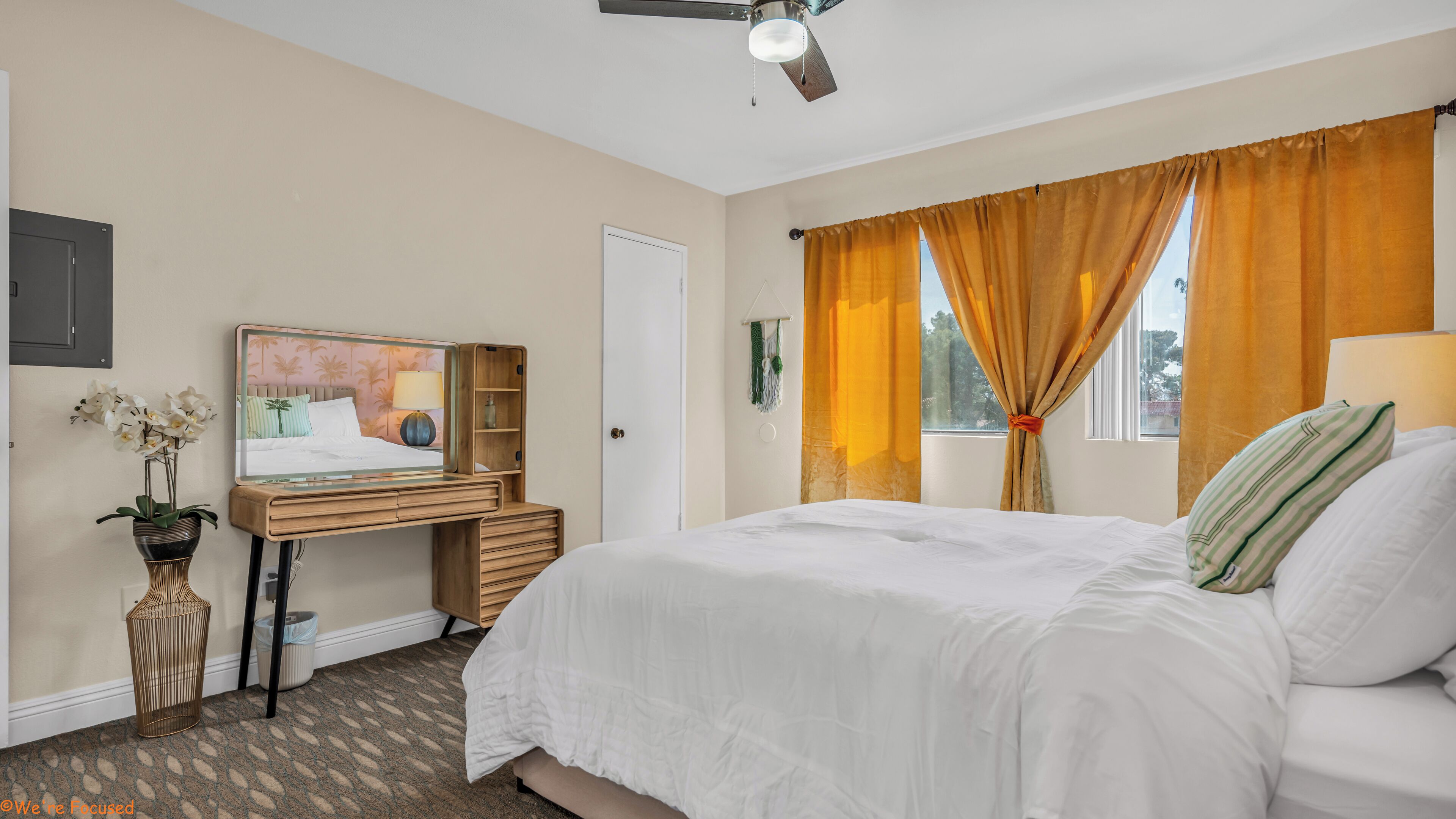 Ein Schlafzimmer mit einem Bett, einem Schreibtisch mit Stuhl, einem Spiegel, einem Fenster mit Vorhängen und einem Deckenventilator.
